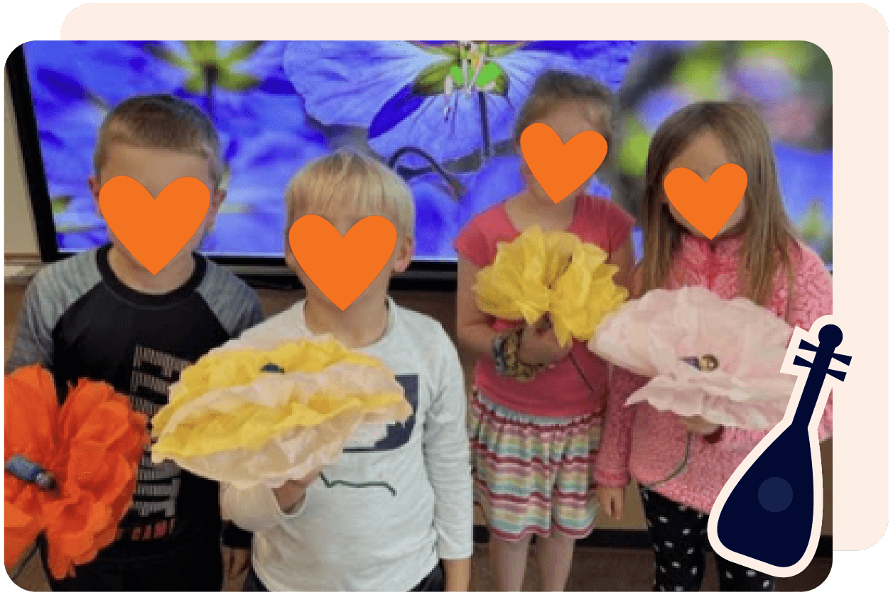 Four children holding large paper flowers in a classroom with a colorful background and an illustration of a stringed instrument in the corner.