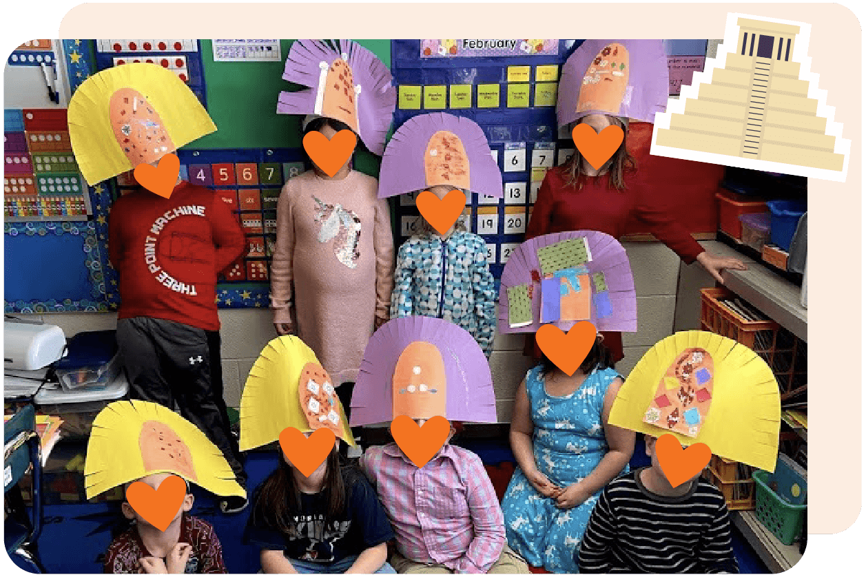 A group of children in a classroom wear colorful paper headdresses with an image of a temple in the corner.