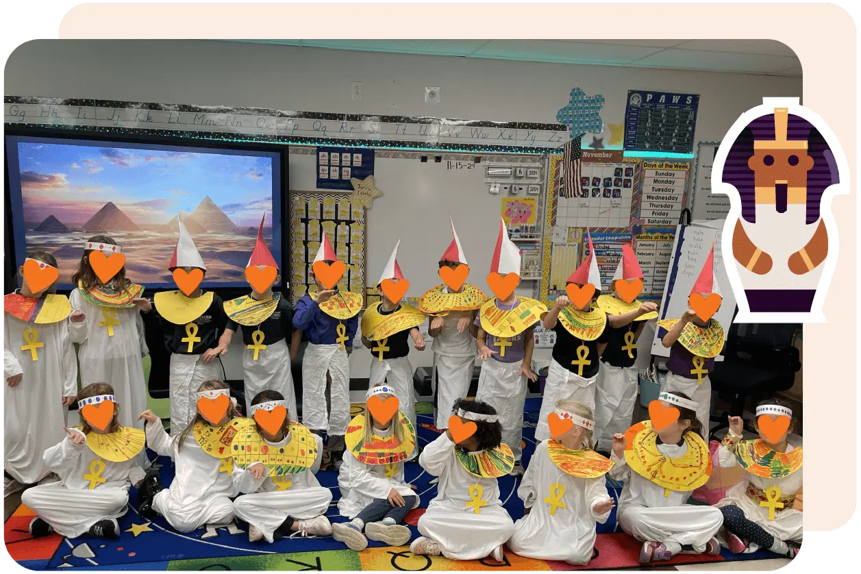 Children dressed in white Egyptian-themed costumes with colorful collars and ankh symbols pose in a classroom decorated with an Egyptian mural and an illustrated pharaoh figure on the wall.