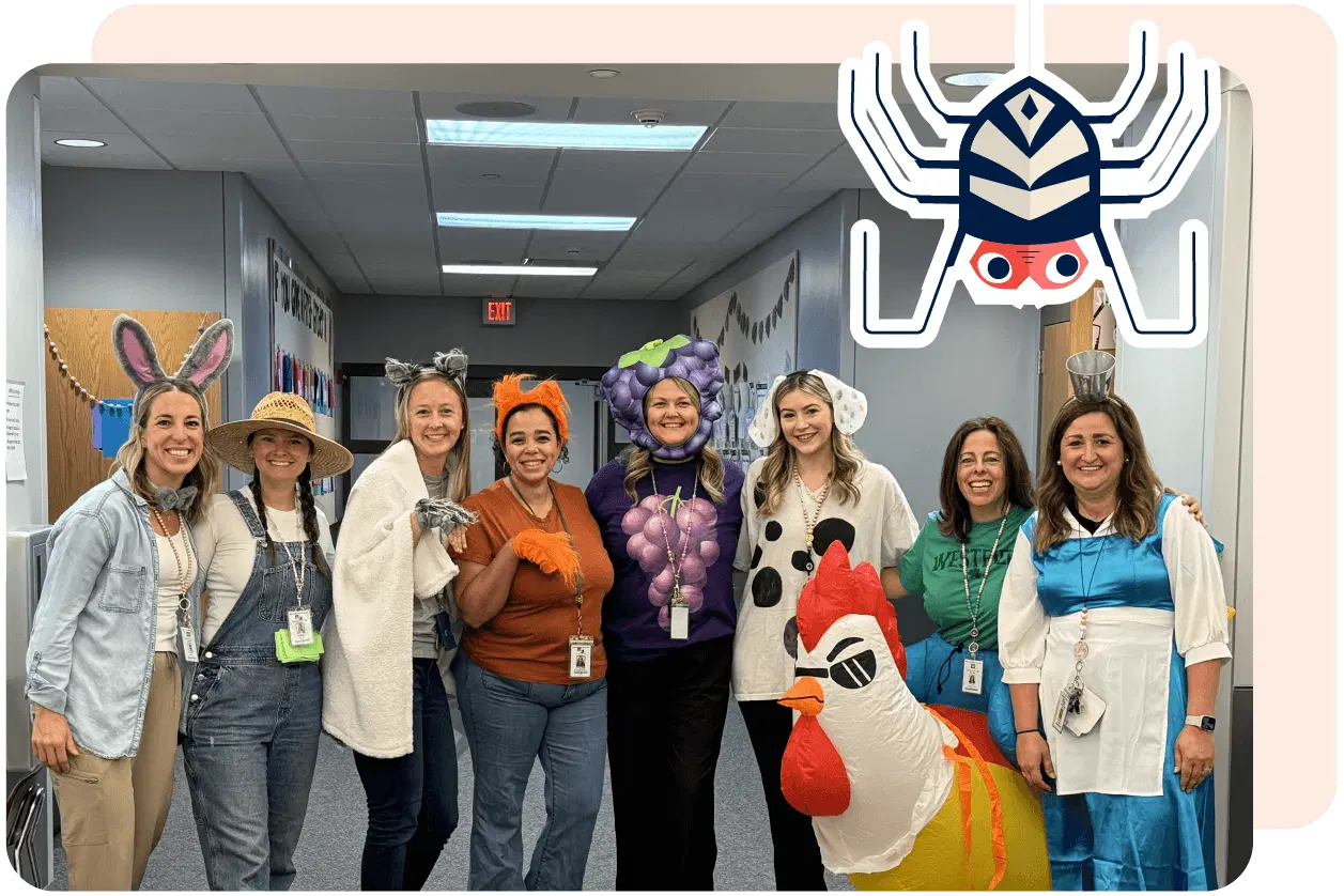 A group of nine people in various creative costumes, including animals and objects, pose together in a hallway decorated with a cartoon spider graphic above them.