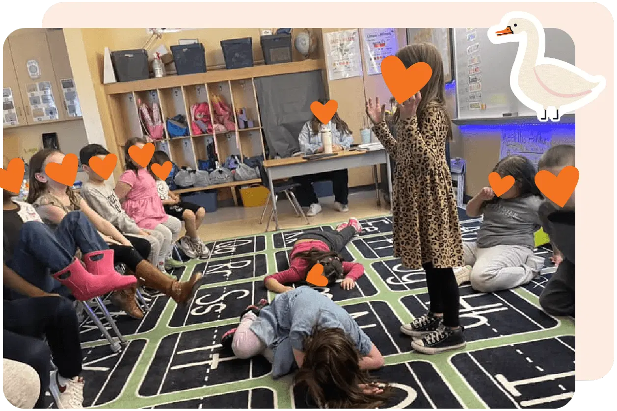 Children in a classroom play a game on a large alphabet rug. Some are sitting in a circle, while one stands and two lie on the floor. A teacher sits nearby at a desk.