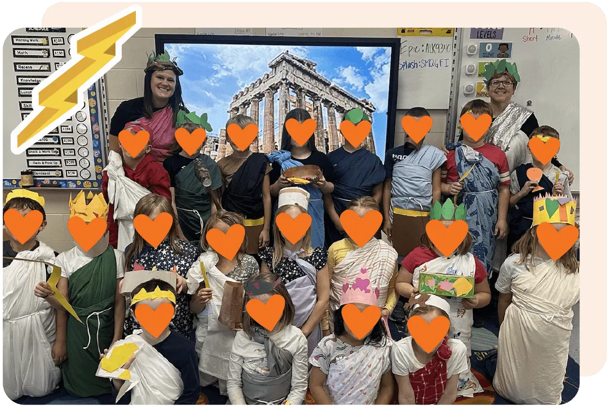 A group of children and two adults dressed in ancient Greek-style clothing poses in front of a screen displaying a Greek temple image.