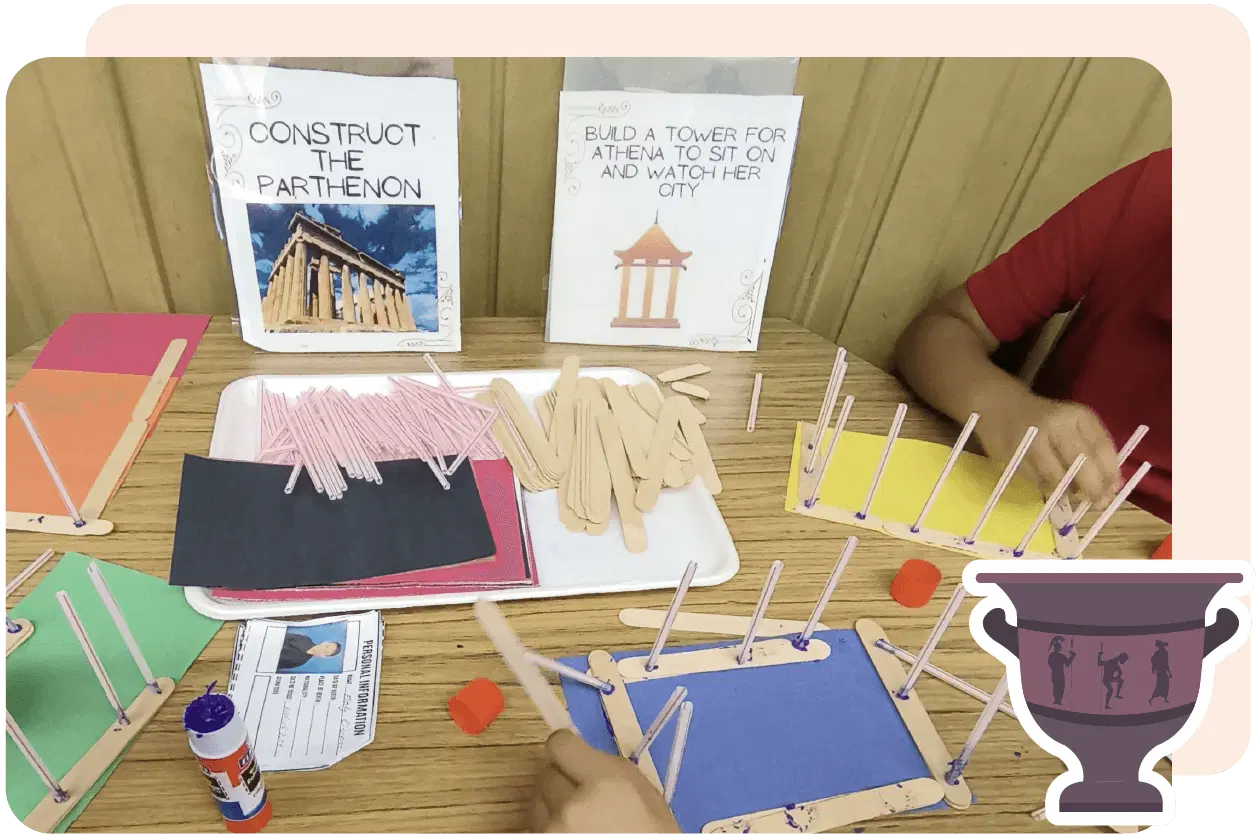 A student builds a model with craft sticks and colored paper. Instructions for constructing the Parthenon are displayed in the background.