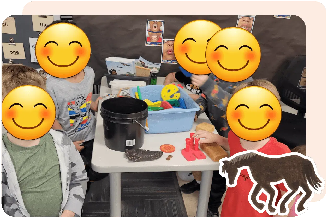 Three children play at a table with toys and buckets. A horse image is on the bottom right. Faces are covered with smiling emojis. Classroom wall with pictures in the background.