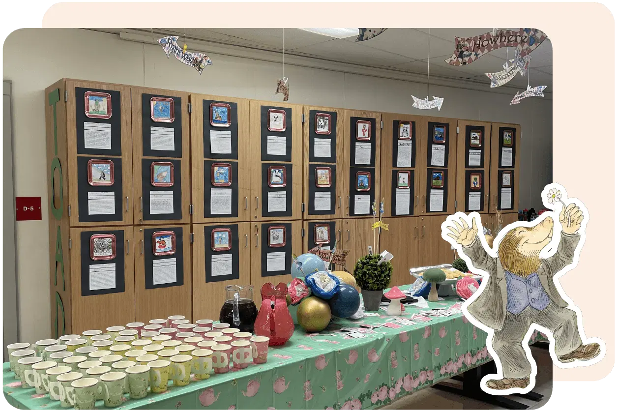 A classroom display with a table set for a tea party, featuring teacups, teapots, and a plant. Wall cabinets hold student artwork and writing. A cartoon character is in the bottom right corner.