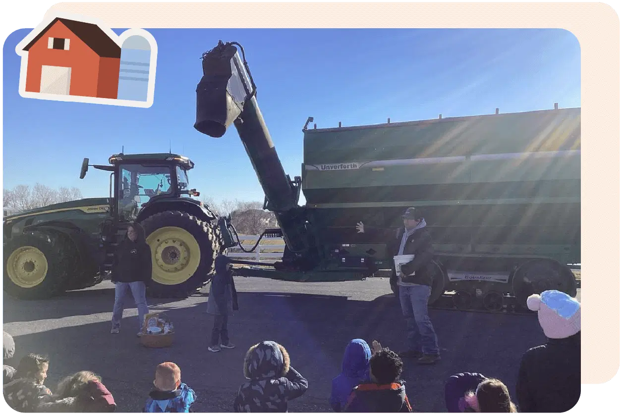 People stand near a large green tractor with an auger wagon, addressing a group of children seated on the ground. An illustrated barn icon is in the top left corner.