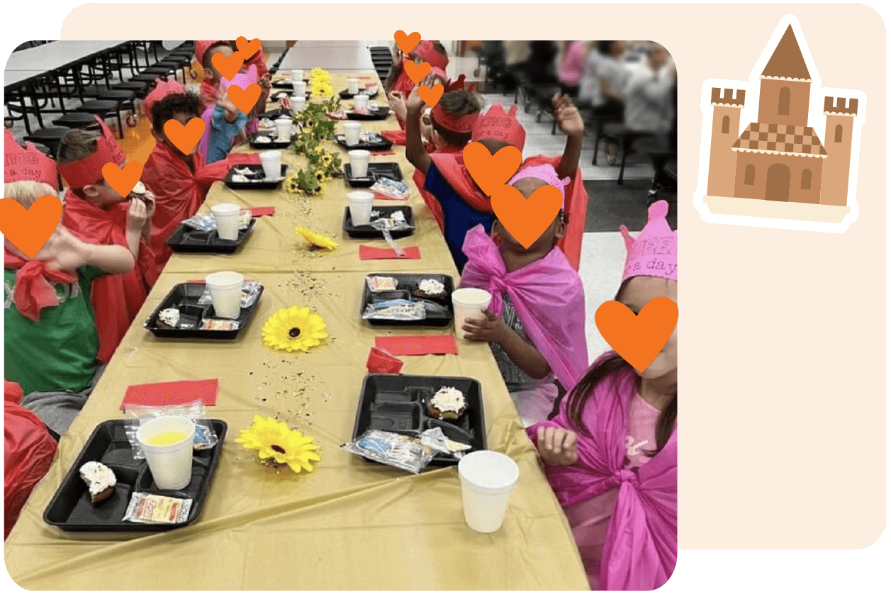 Children wearing red and purple paper crowns and capes sit at a long table decorated with sunflowers, having lunch.