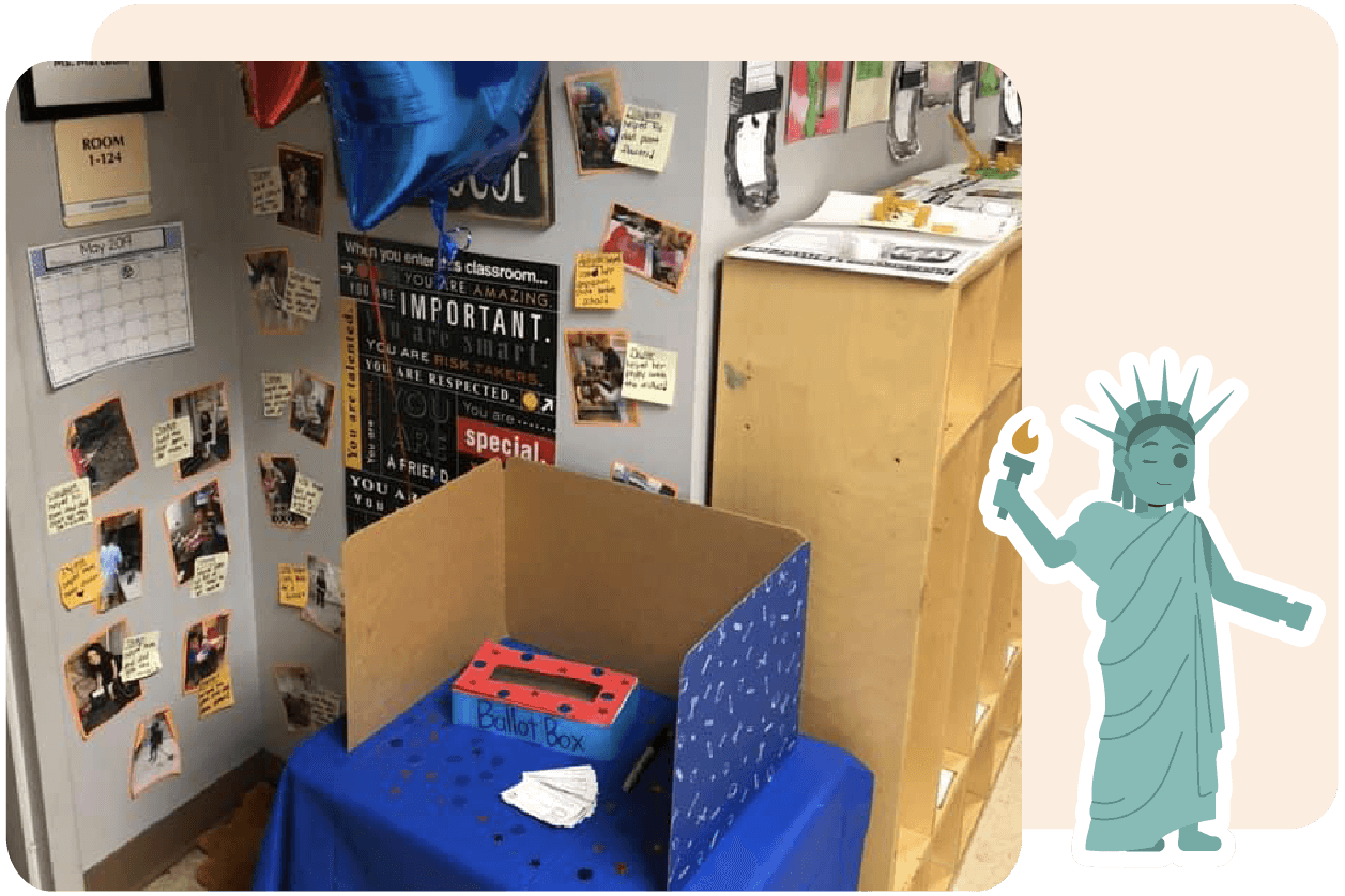 A ballot box is placed on a table with blue fabric. The background displays photos, a calendar, and diverse posters on a classroom wall.