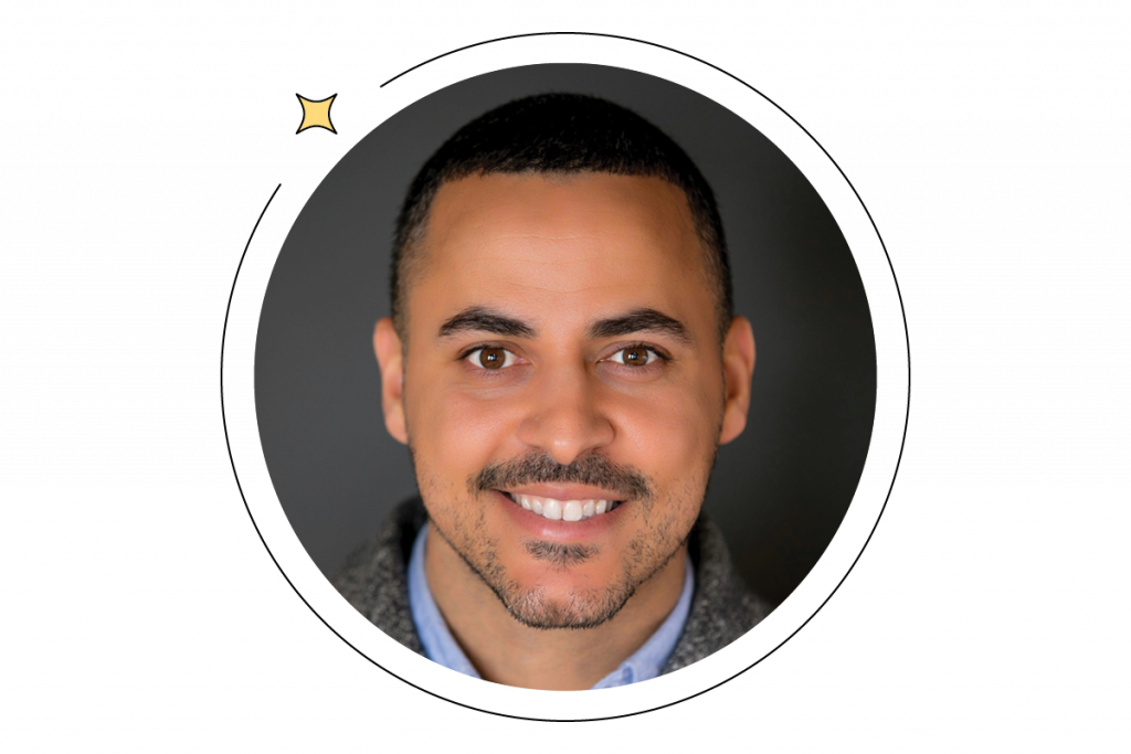 Smiling person with short hair and a beard, wearing a collared shirt and sweater, against a dark background. Small star icon on the top left of the circular frame.