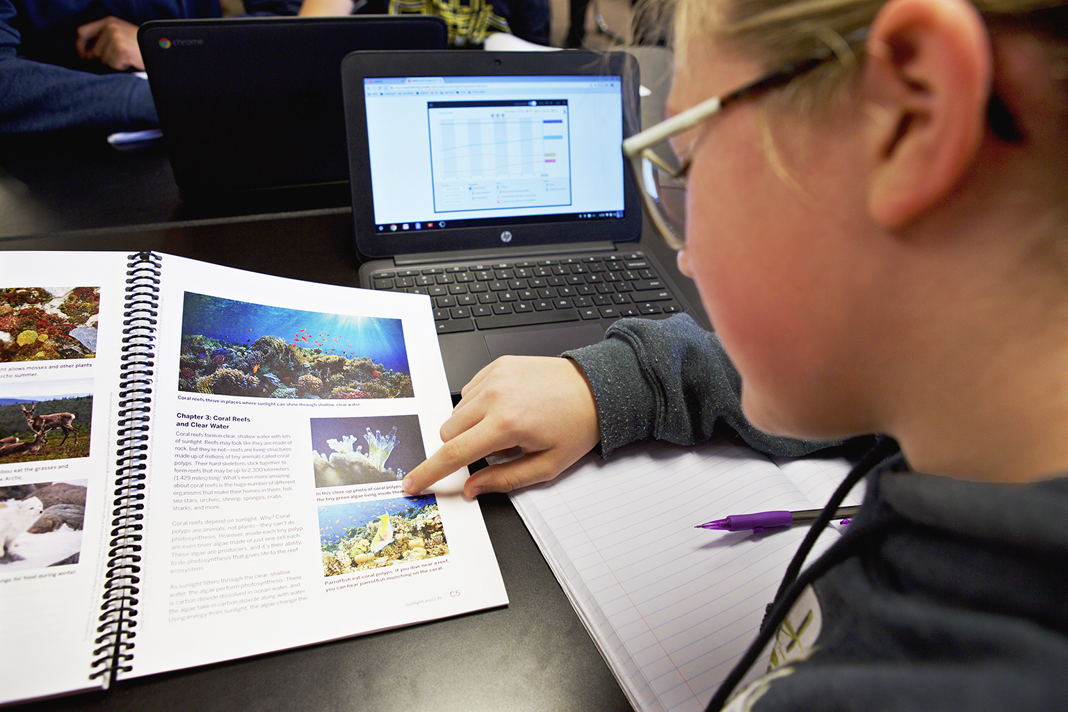 Student reading a textbook about coral reefs, pointing to a page, with a pen and notebook nearby. A laptop displaying a chart is open on the desk.
