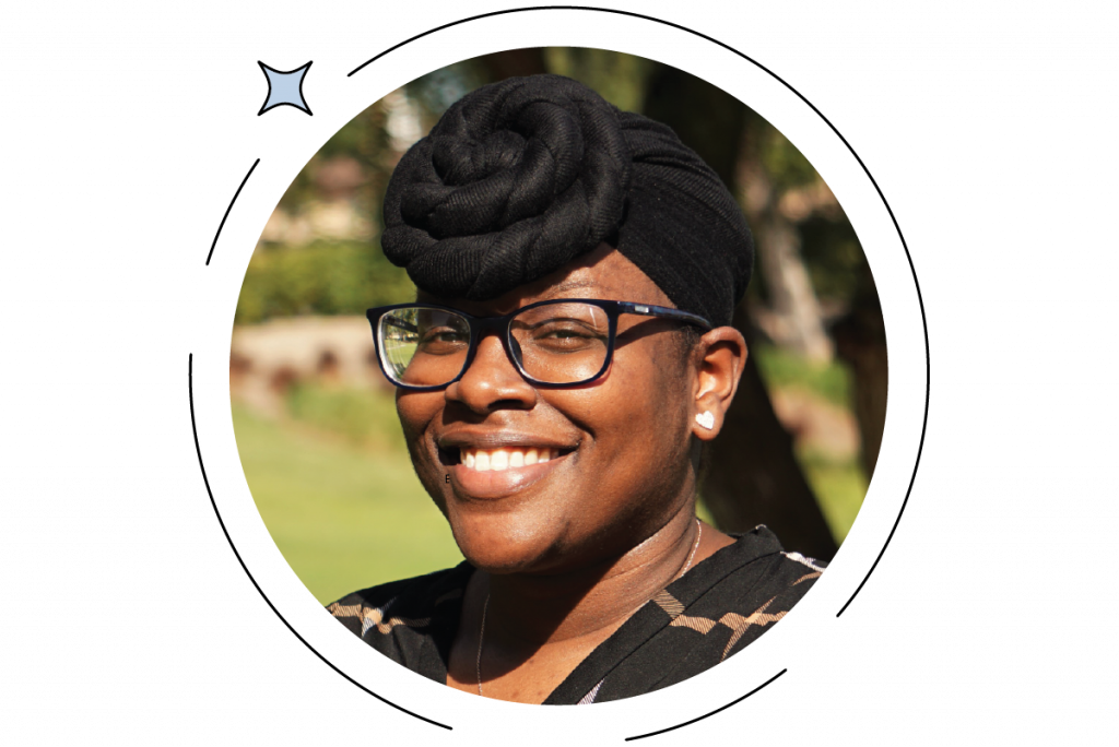Person with glasses smiling, wearing a black headwrap and earrings, outdoors with trees and sunlight in the background.