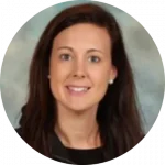 Portrait of a woman with long dark hair, smiling warmly, wearing a black top. She is posed against a neutral, softly blurred background, embodying the confidence and wisdom often seen in educators passionate about early literacy and the science of reading curriculum.