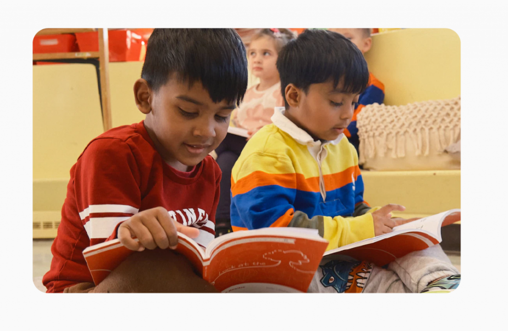 Two children are sitting on the floor, reading books. One wears a red shirt, the other a colorful sweater. Other children are seated in the background.