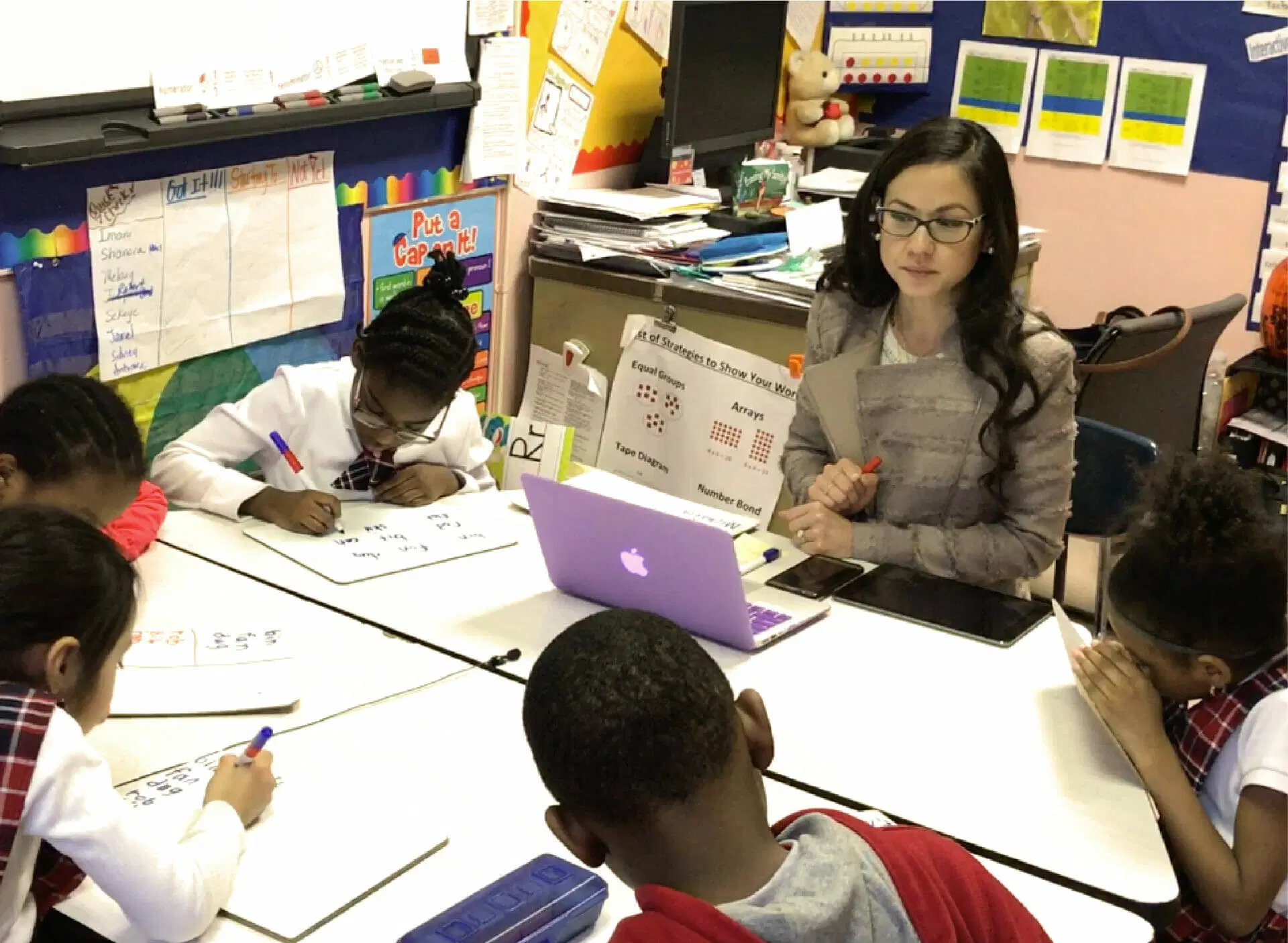 A teacher with glasses sits at a table with five students, aiding them in writing exercises as part of mCLASS Intervention. A pink laptop is open on the table, while educational posters adorn the classroom walls.