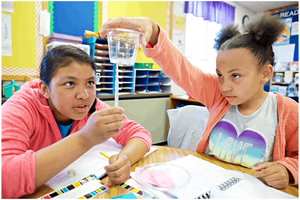 Two students conduct a water experiment in a classroom. One holds a cup while the other observes. Books and papers are on the table.