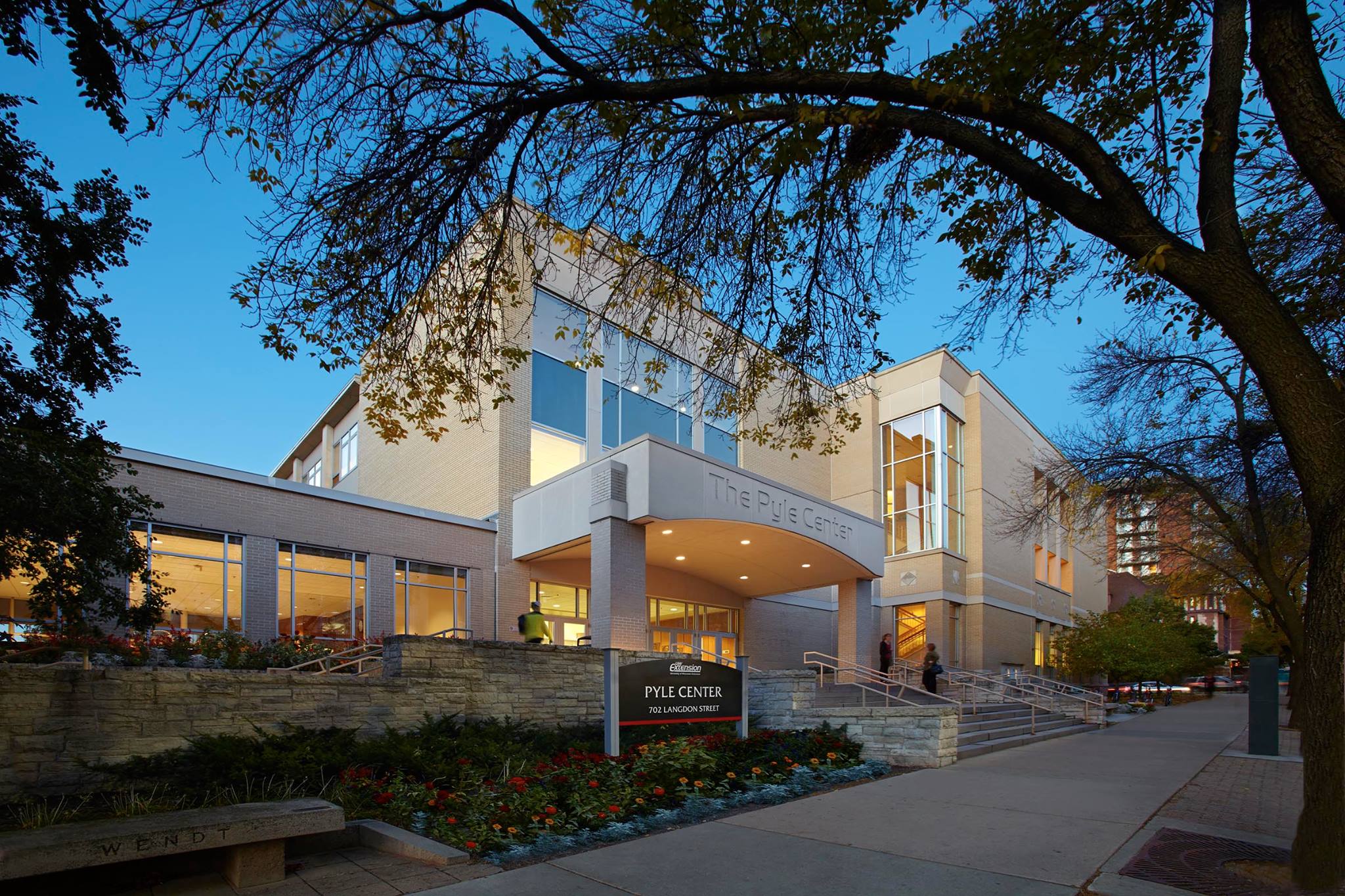 Exterior of the Pyle Center at dusk, featuring modern architecture, large windows, and a sign in front. Leafy trees frame the scene.