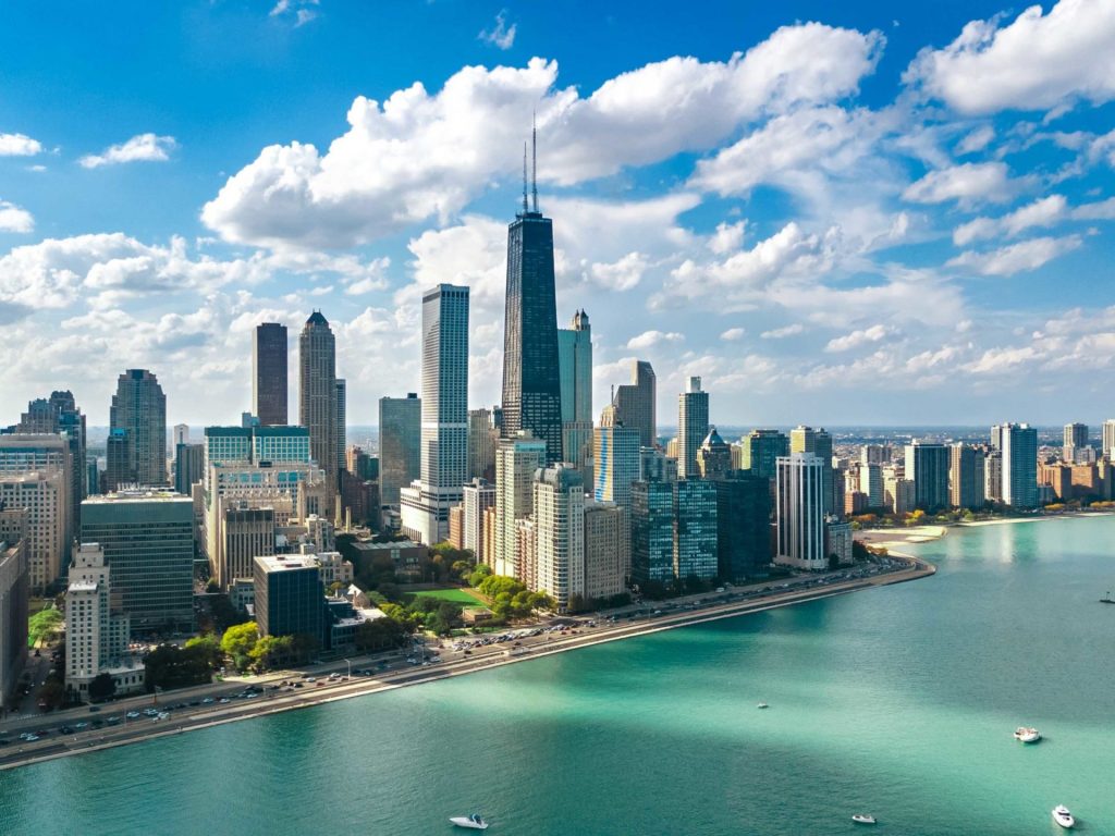Aerial view of a city skyline with tall skyscrapers, a waterfront, and a road along the shore. The sky is partly cloudy, and the water is a light turquoise.