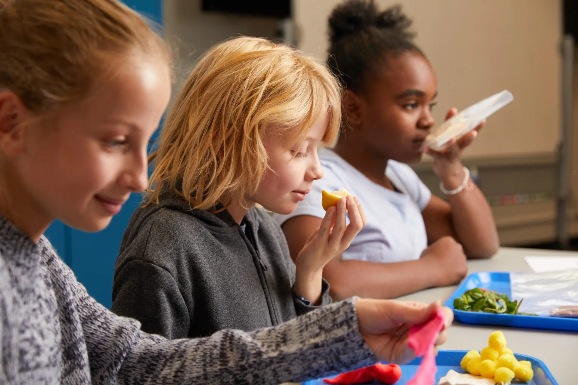 Three children sit at a table smelling and examining pieces of food on blue trays, participating in a sensory activity.