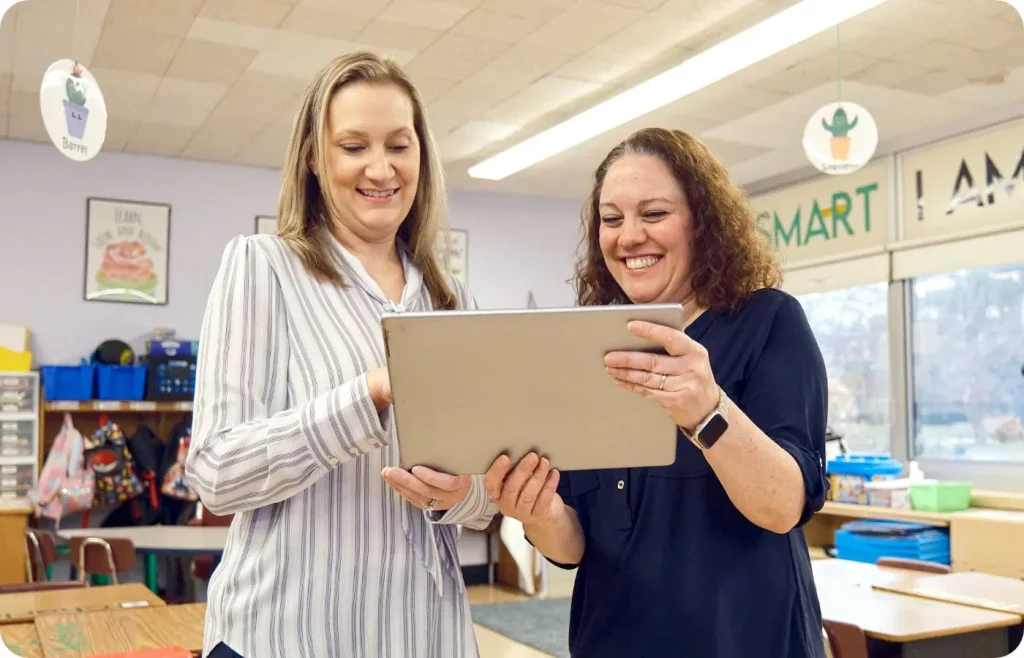 Two women stand in a classroom, smiling as they look at a laptop together—sharing teaching tips and supporting each other in the education community.