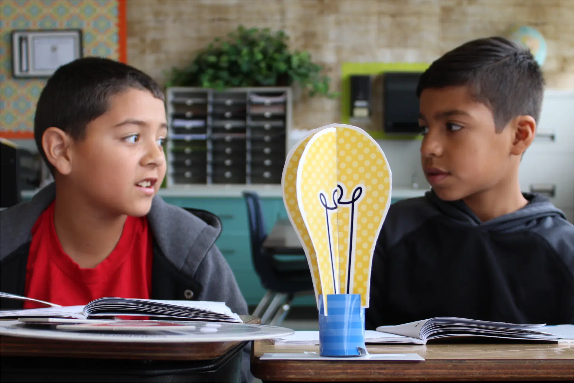Two boys sit at desks in a classroom, talking to each other. An upright paper cutout of a yellow light bulb is placed between their open books.