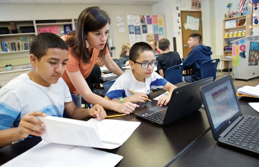 Teacher assisting two students working on laptops in a classroom; other students are in the background.