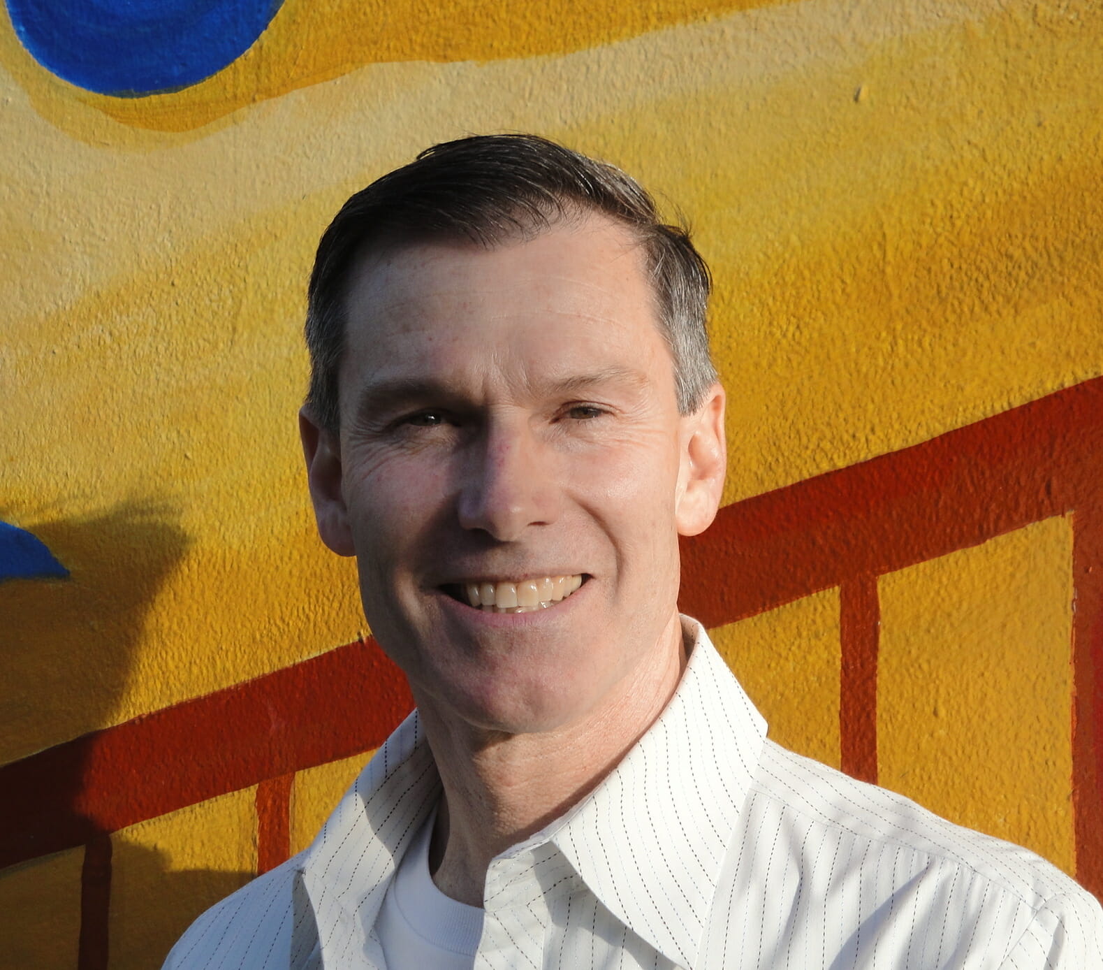 Person smiling in front of a colorful mural with yellow and red geometric shapes.