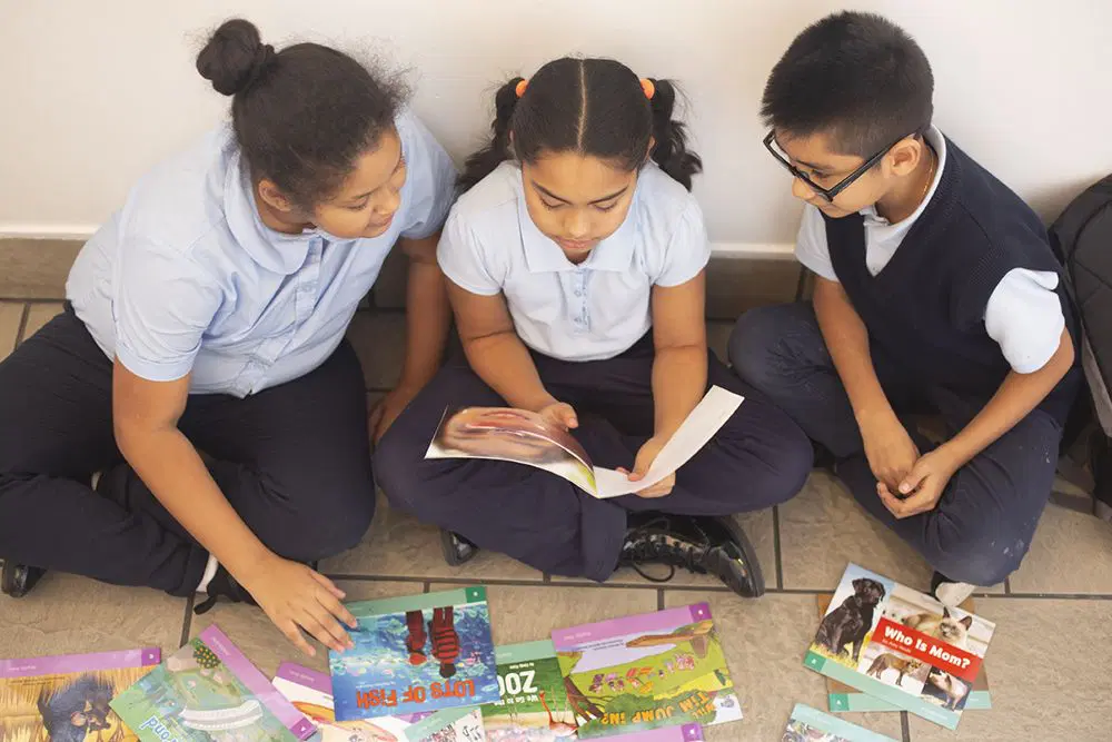 Three children in school uniforms sit on the floor reading books together, with several colorful books spread out in front of them.