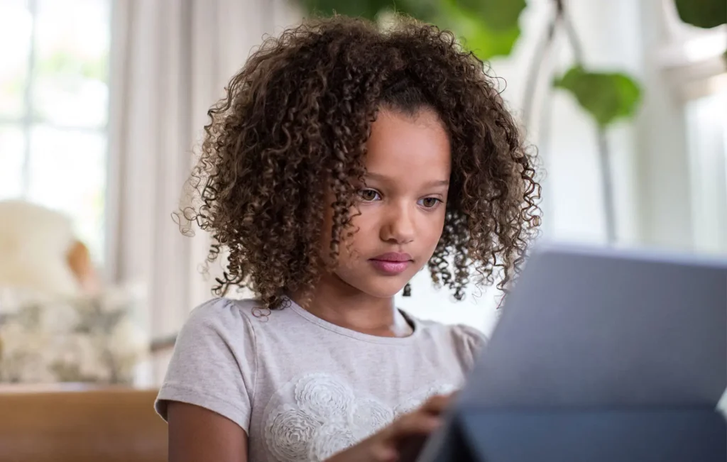 Young girl with curly hair sits indoors, looking at a tablet device with a focused expression.