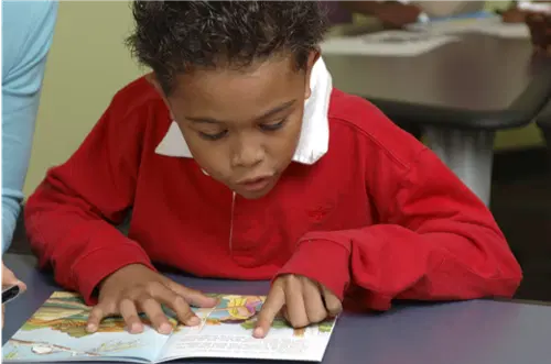 A young boy in a red shirt sits at a table, reading a picture book and pointing at the text with his finger.