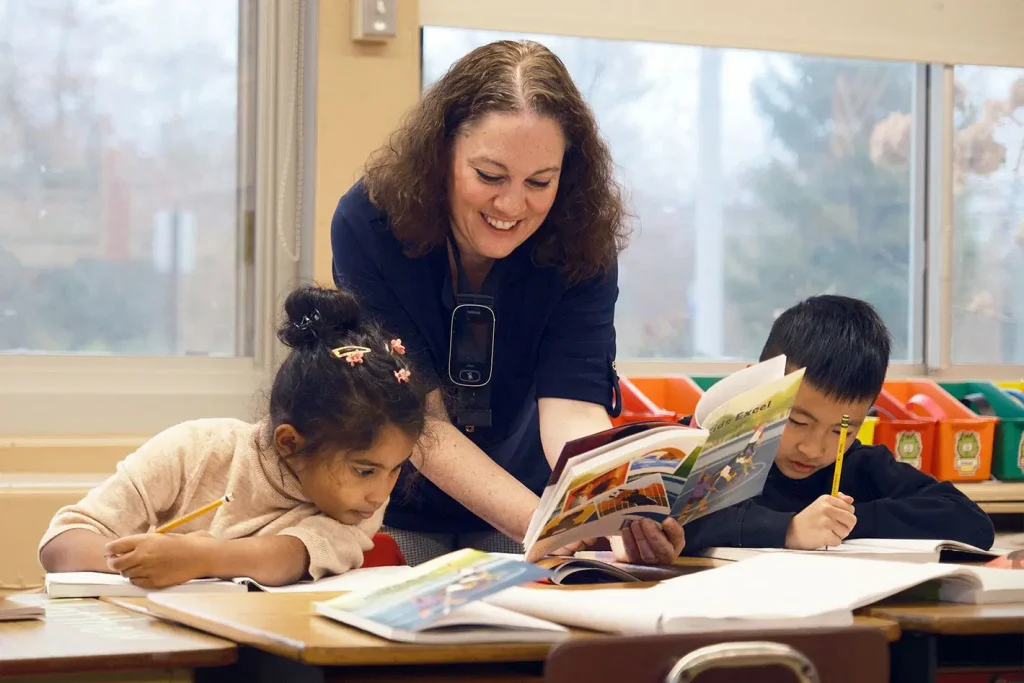 A teacher helps two young students with their reading and writing at a classroom desk.
