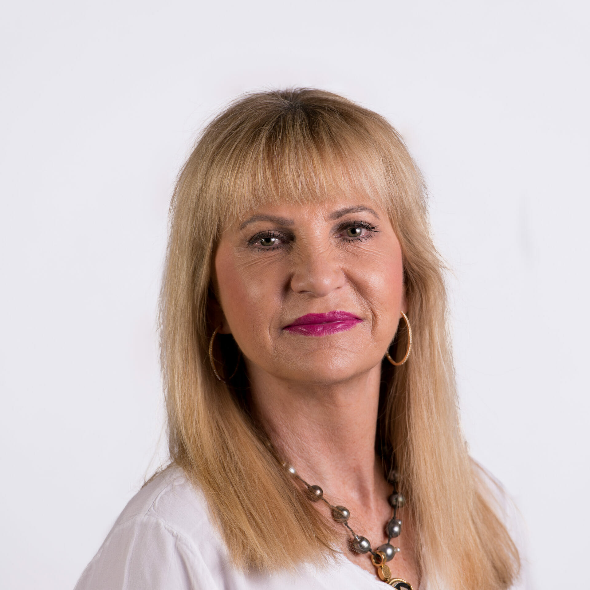 A woman with medium-length blonde hair and hoop earrings, wearing a white shirt and a beaded necklace, is looking directly at the camera against a plain background.