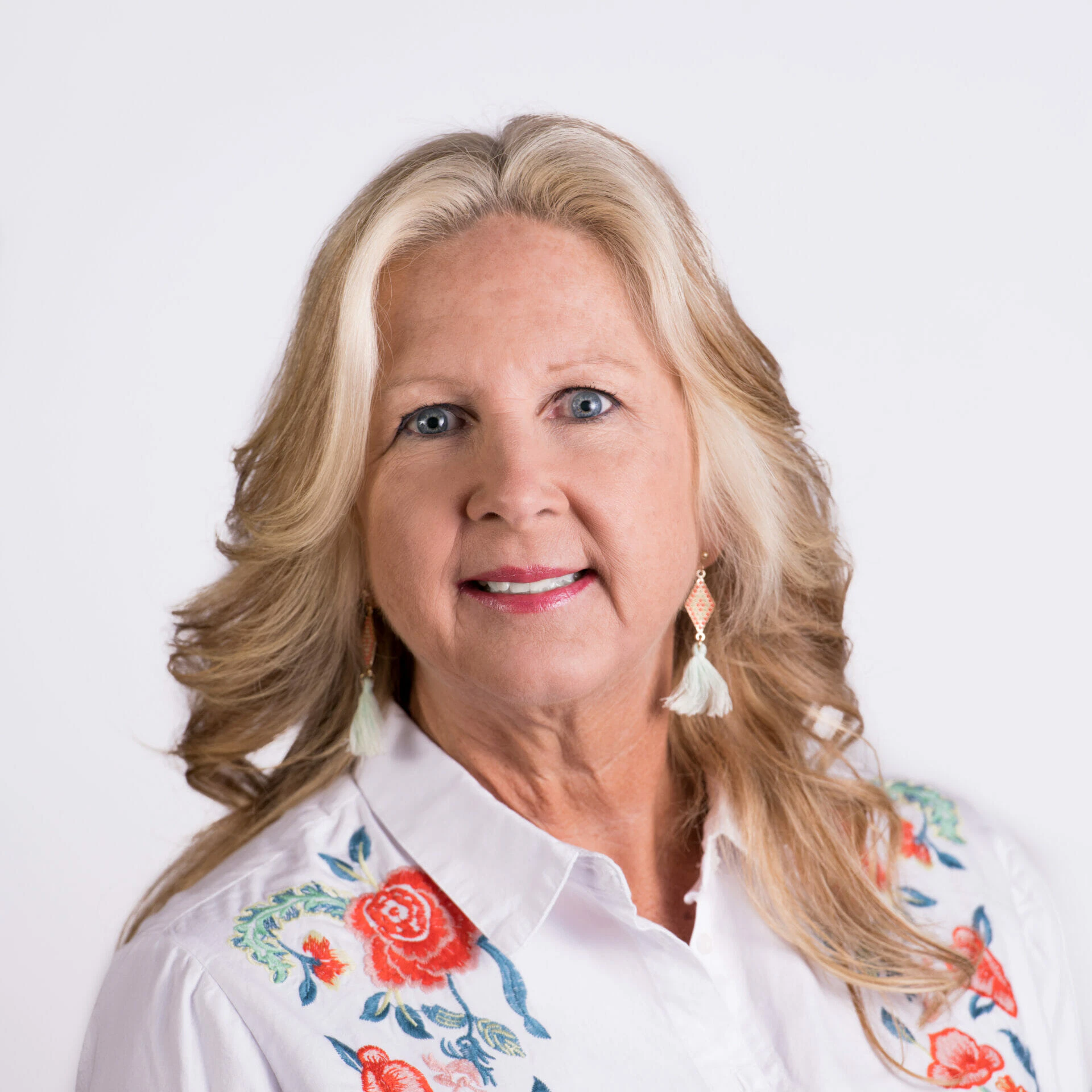 A woman with blonde hair wearing a white blouse with floral embroidery, posing against a plain white background.