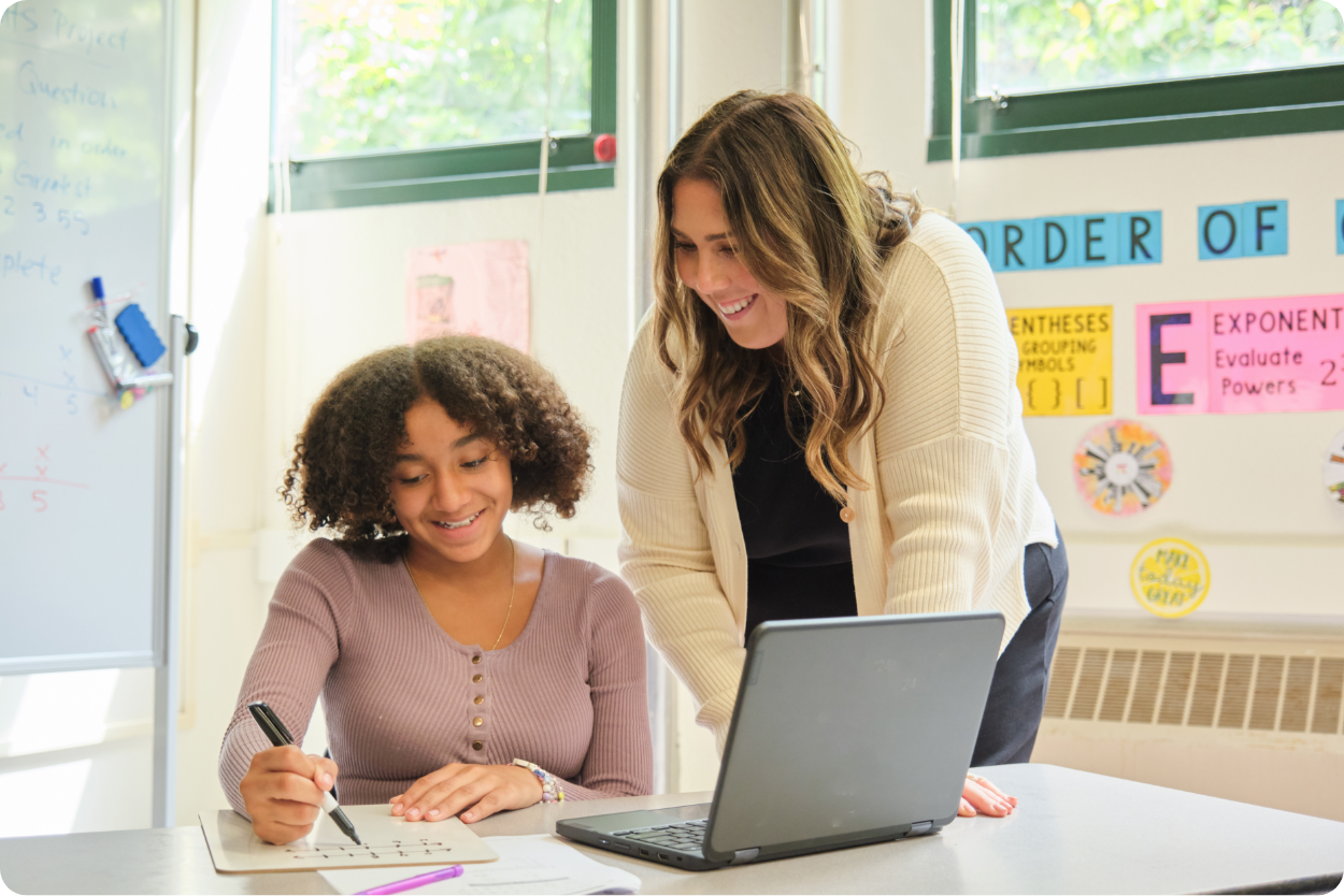 A student writes in a notebook at a desk with a laptop, as her awesome teacher stands beside her. Both appear engaged in conversation about her academic struggles during ELA class.