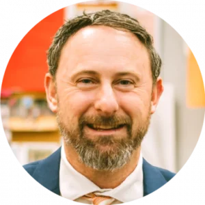 A middle-aged man with short brown hair and a beard, wearing a navy suit, white shirt, and striped tie, smiles in an indoor setting, radiating confidence often seen in champions of literacy success.