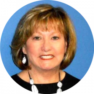 A woman with light brown hair, wearing a black top, white earrings, and a necklace, smiles in front of a blue background, embodying literacy success with her confident and approachable presence.