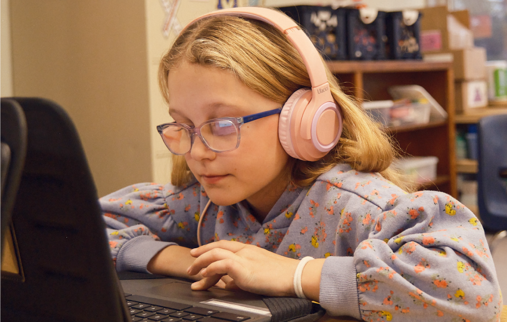 Young girl wearing glasses and pink headphones looks at a laptop screen while sitting at a desk in a classroom, participating in a Remote Learning Language Arts Program.