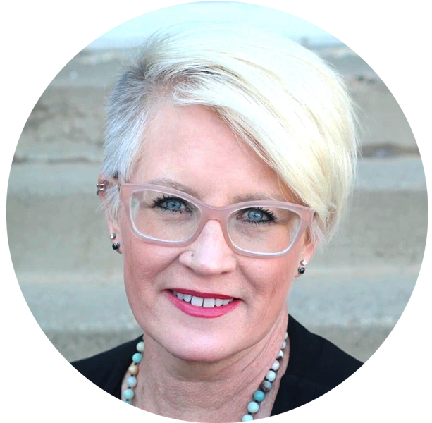 A woman with short, light blonde hair, wearing clear glasses, a black top, and a beaded necklace, smiles at the camera in front of a blurred outdoor background.