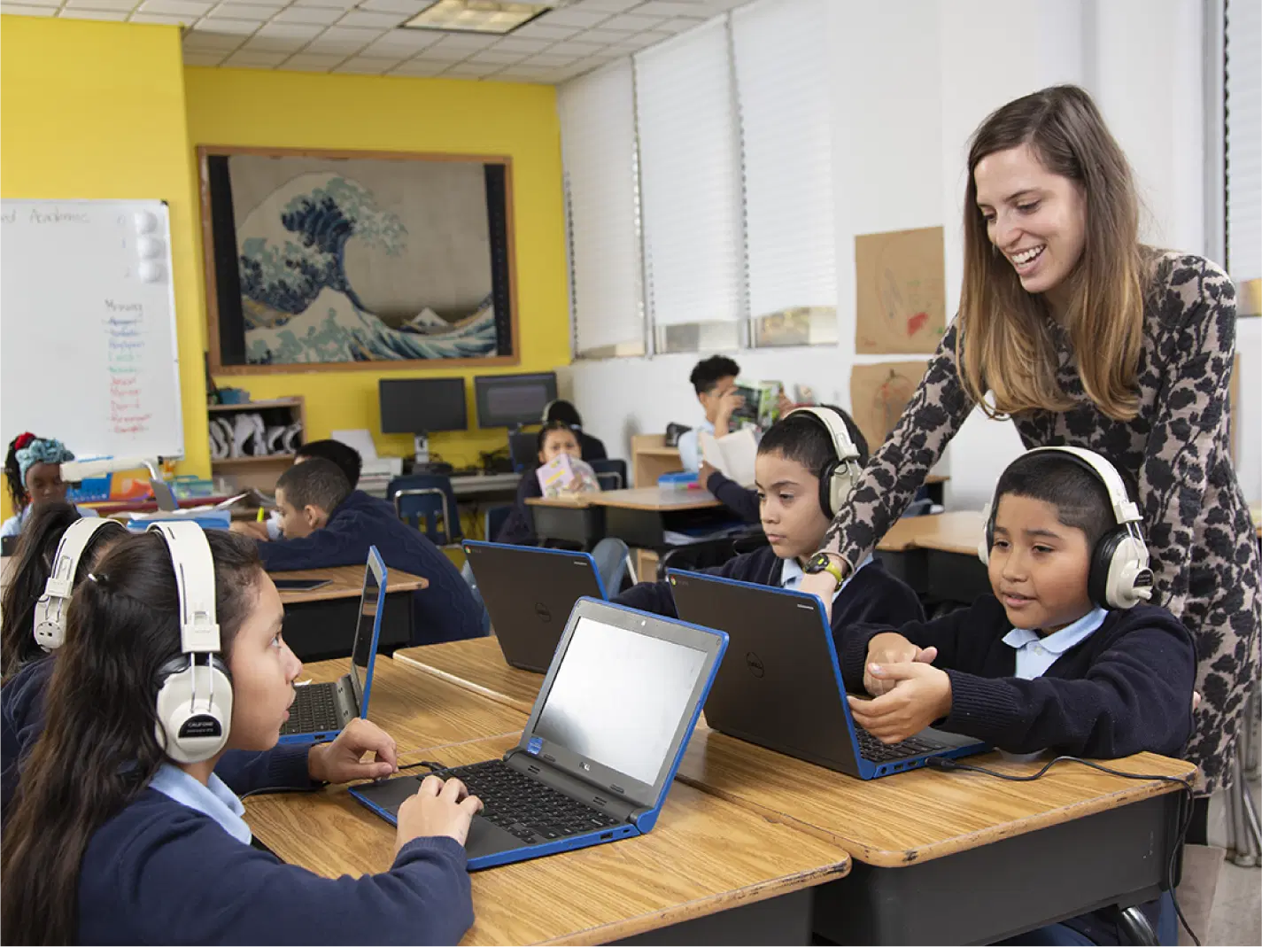A teacher stands next to students wearing headphones and working on laptops in a classroom setting. Other students are visible in the background.