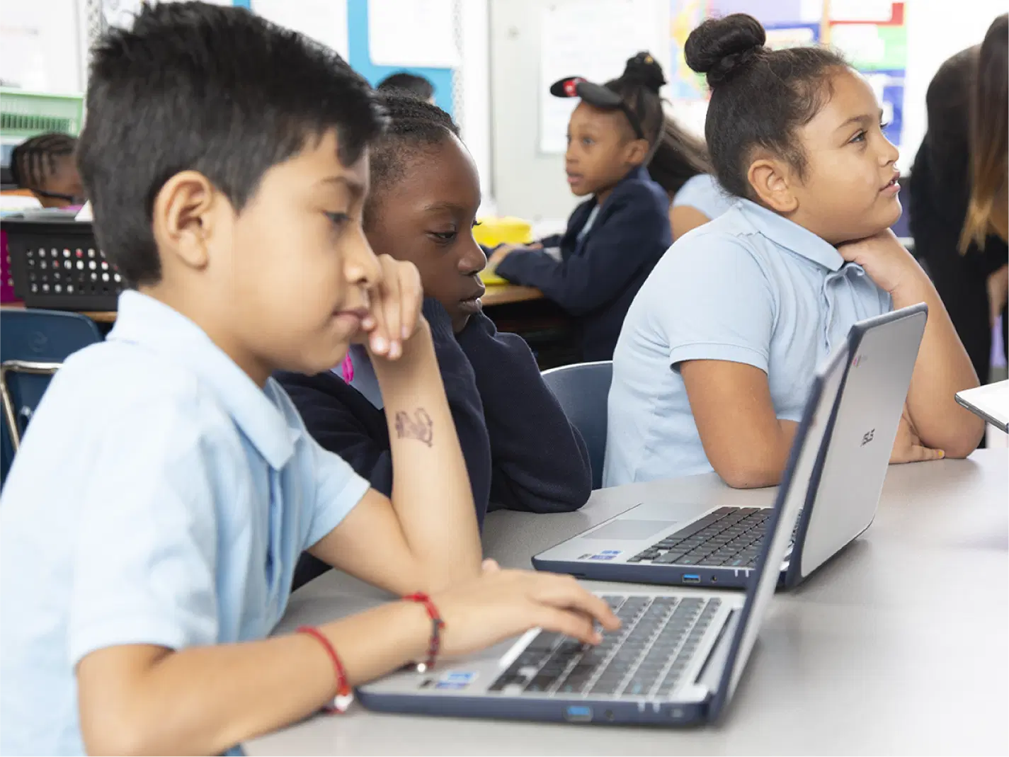 Three children sit at a classroom table, each using a laptop, with other students visible in the background.