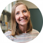 A woman with straight, shoulder-length blonde hair smiles at the camera, wearing a striped sweater and hoop earrings. Shelves with books are visible in the background.