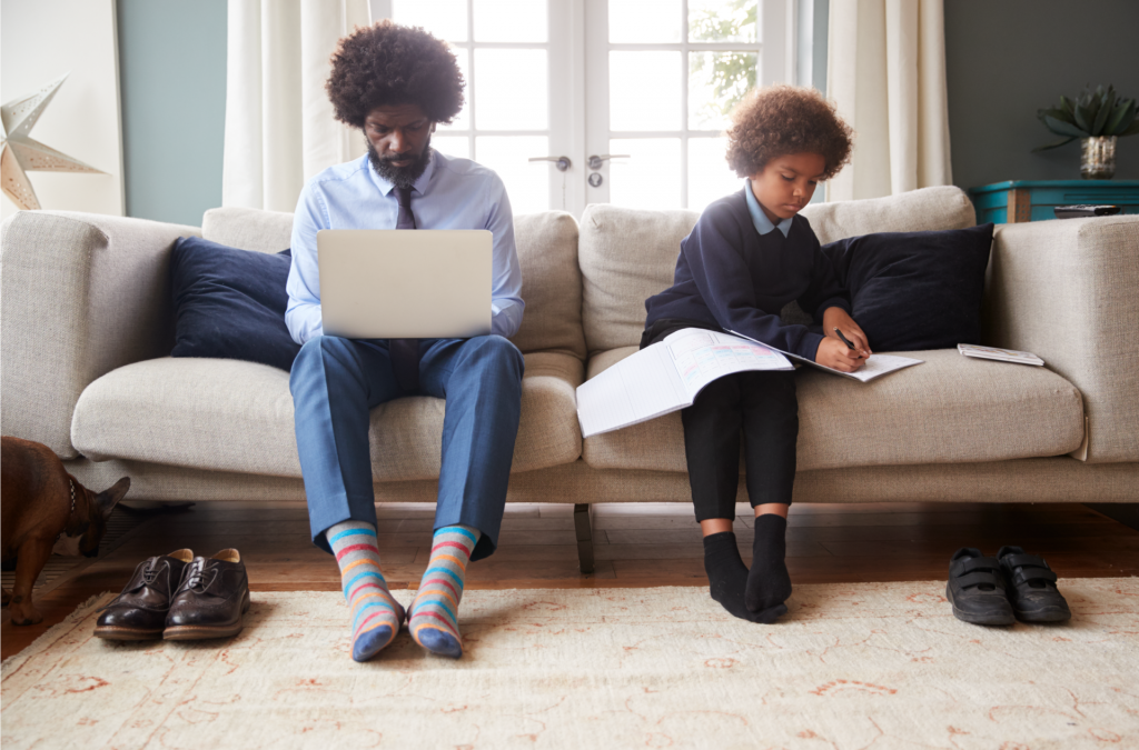 An adult and a child sit on a sofa. The adult works on a laptop, while the child writes in a notebook. Two pairs of shoes are on the floor, and a dog is visible on the left side.