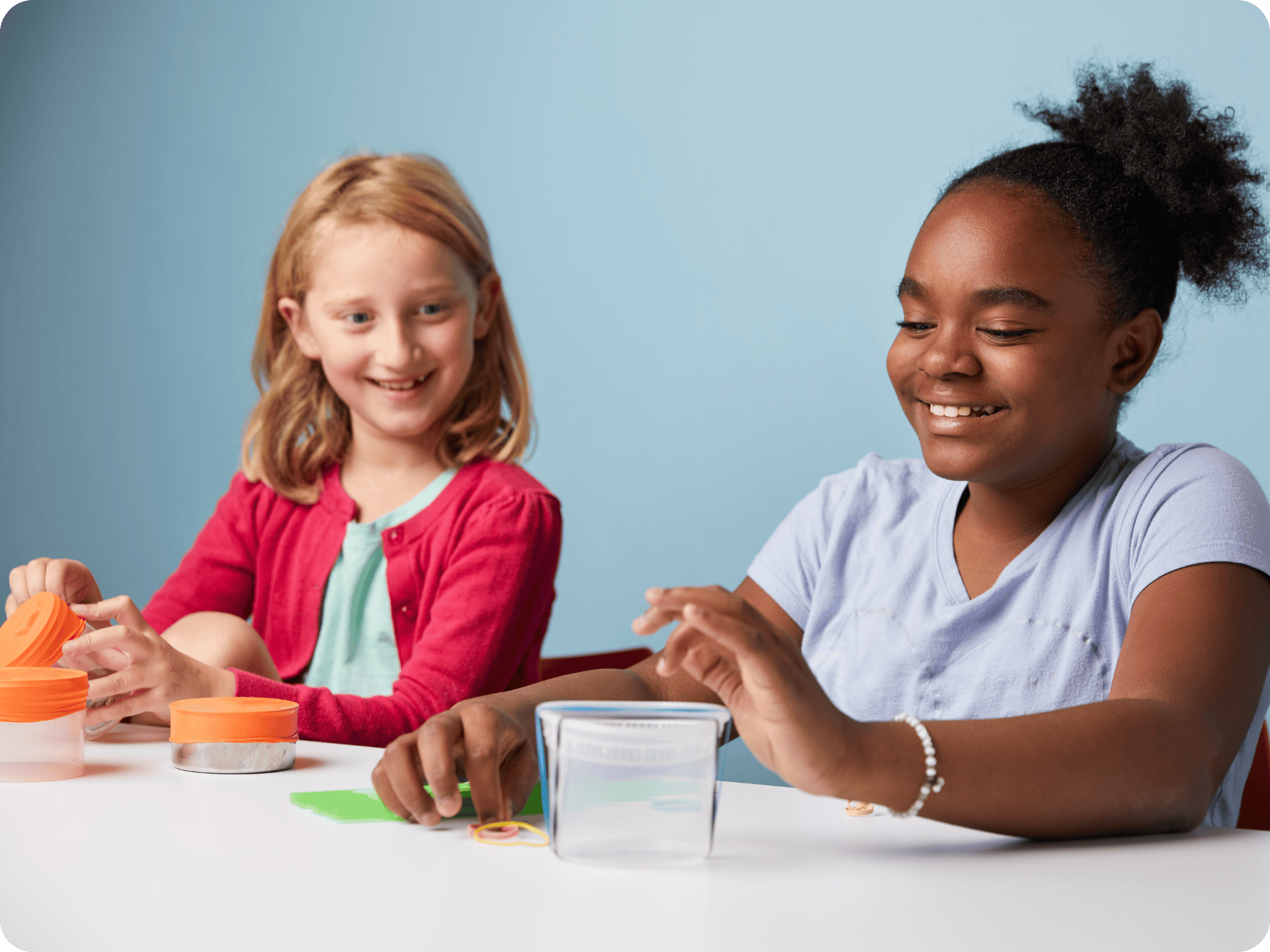 Two girls sit at a table smiling and playing with colorful modeling clay and clear containers against a light blue background.