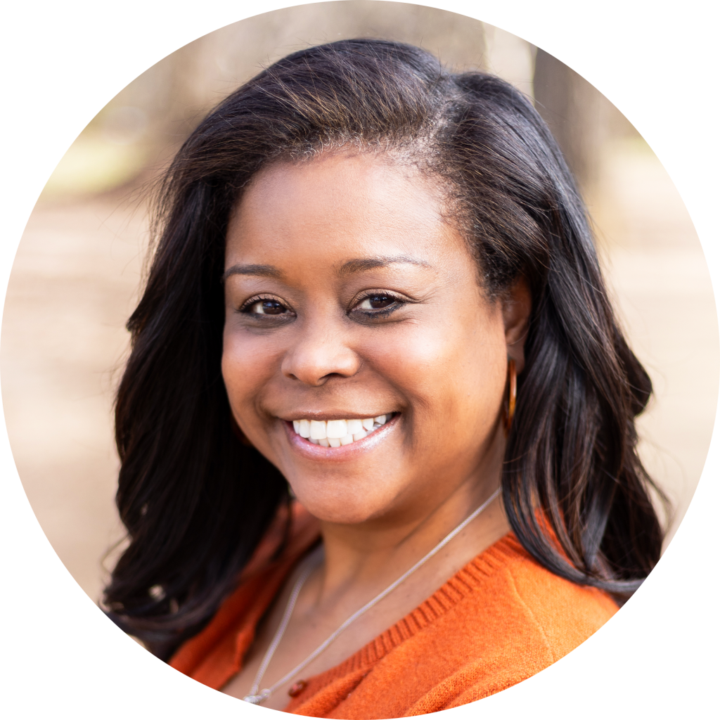 A woman with long dark hair and a bright smile, wearing an orange sweater and a necklace, poses outdoors with blurred trees in the background, radiating confidence often seen in literacy education and academic language development.