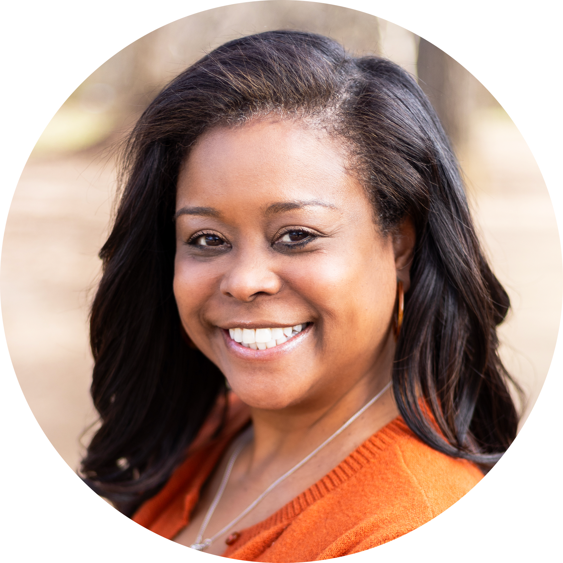 A woman with long dark hair and a bright smile, wearing an orange sweater and a necklace, poses outdoors with blurred trees in the background, radiating confidence often seen in literacy education and academic language development.