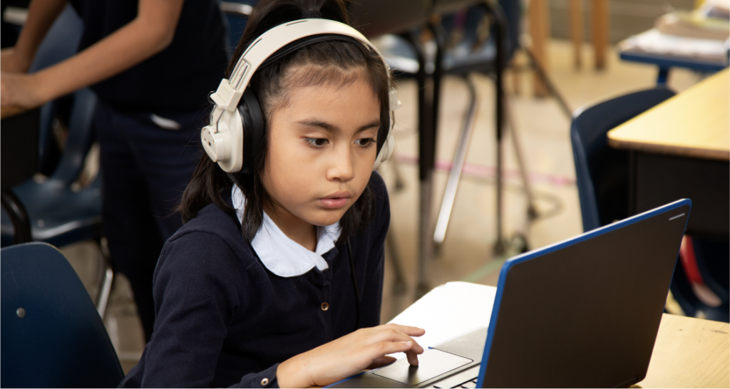 A student wearing headphones sits at a desk, looking at a laptop screen and using the trackpad in a classroom setting.