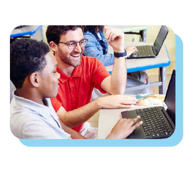 A teacher in a red shirt sits beside a student, both looking at a laptop screen in a classroom setting with other students visible in the background.