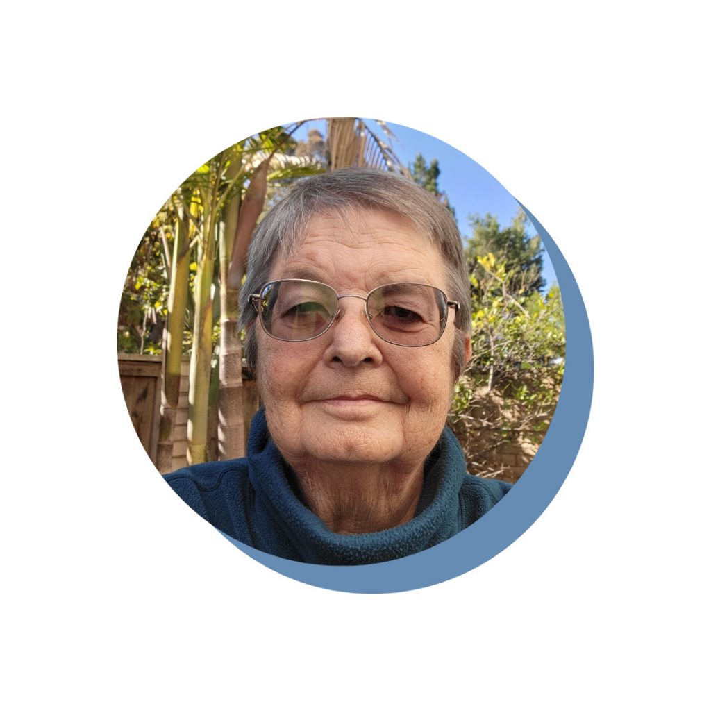 An older person with short gray hair and glasses, wearing a blue sweater, is outdoors with greenery and a fence in the background.