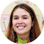 A young woman with long brown hair smiles at the camera, wearing hoop earrings and a lanyard, with colorful posters on the wall behind her.