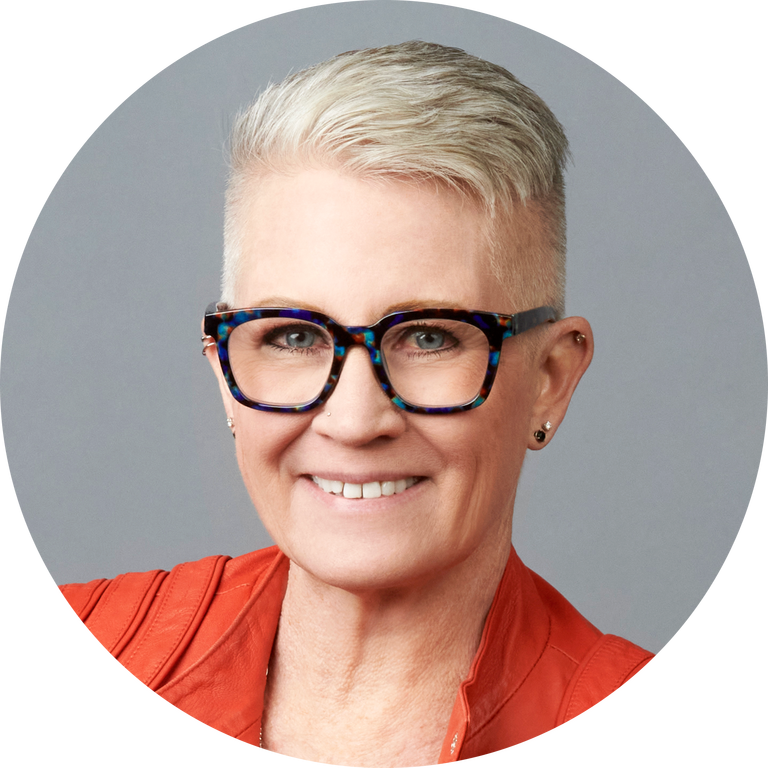 Person with short blonde hair, glasses, and earrings, wearing an orange jacket, smiling in front of a plain gray background—committed to literacy education and fostering background knowledge for all learners.