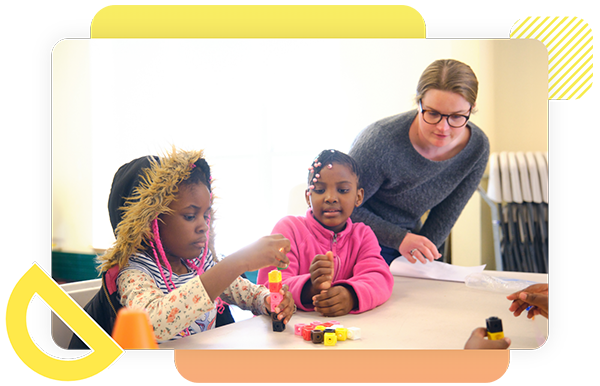 Two young girls build with colorful blocks at a table while an adult woman observes and assists them in a classroom setting.